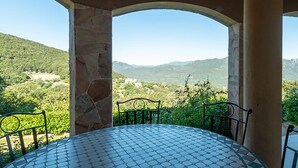 Uma área de estar coberta ao ar livre, com um pilar de pedra e cadeiras de metal, com vista para uma paisagem montanhosa.