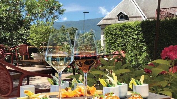 Un espacio de comedor al aire libre con una mesa dispuesta para dos, que incluye una copa de vino, aperitivos y vistas a una casa y montañas.