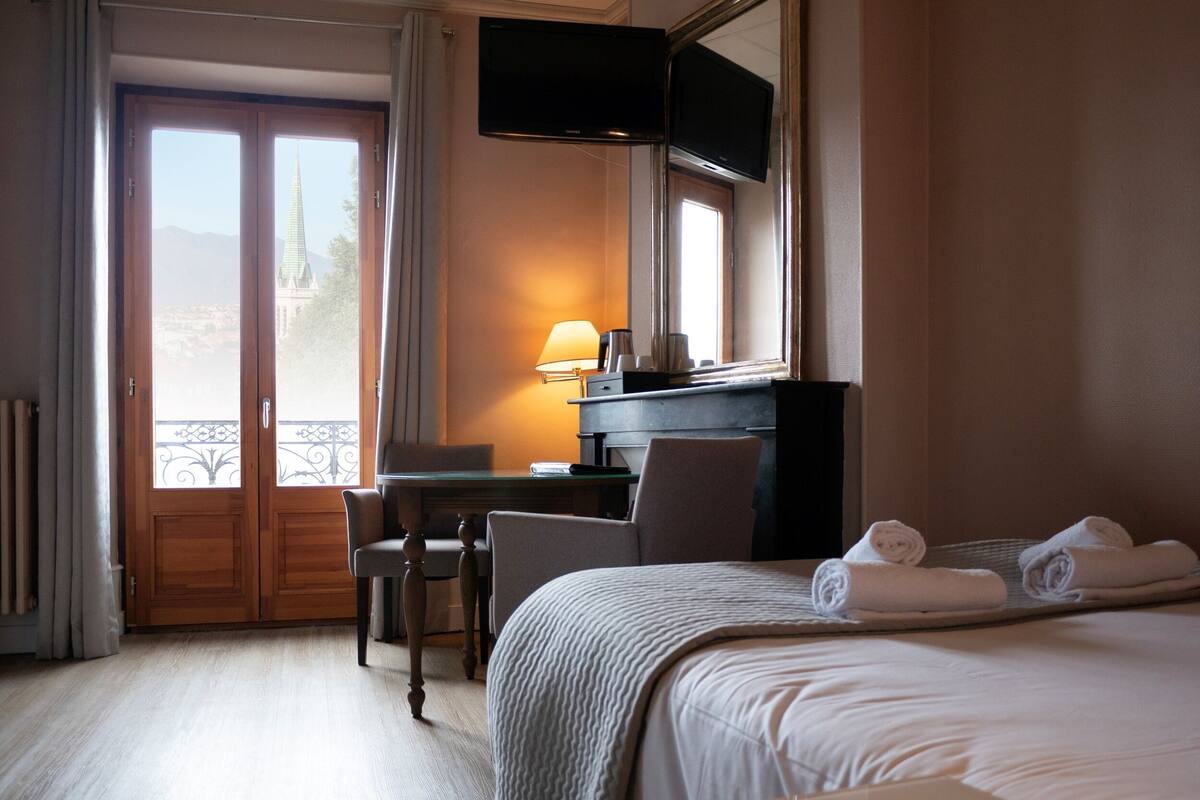 Habitación de hotel con cama, escritorio, silla, televisor y balcón con vistas.