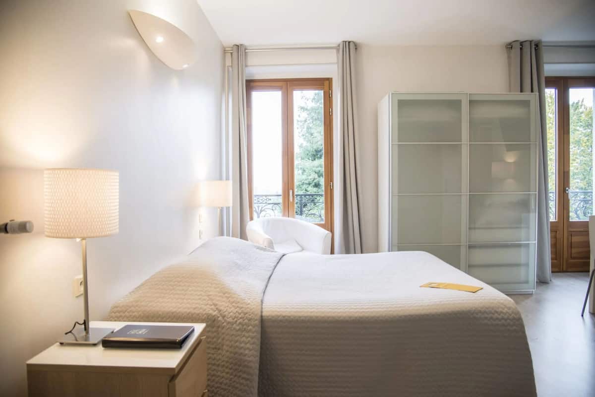 Una habitación de hotel con cama, lámparas de noche, una silla, armario y ventana con cortinas.