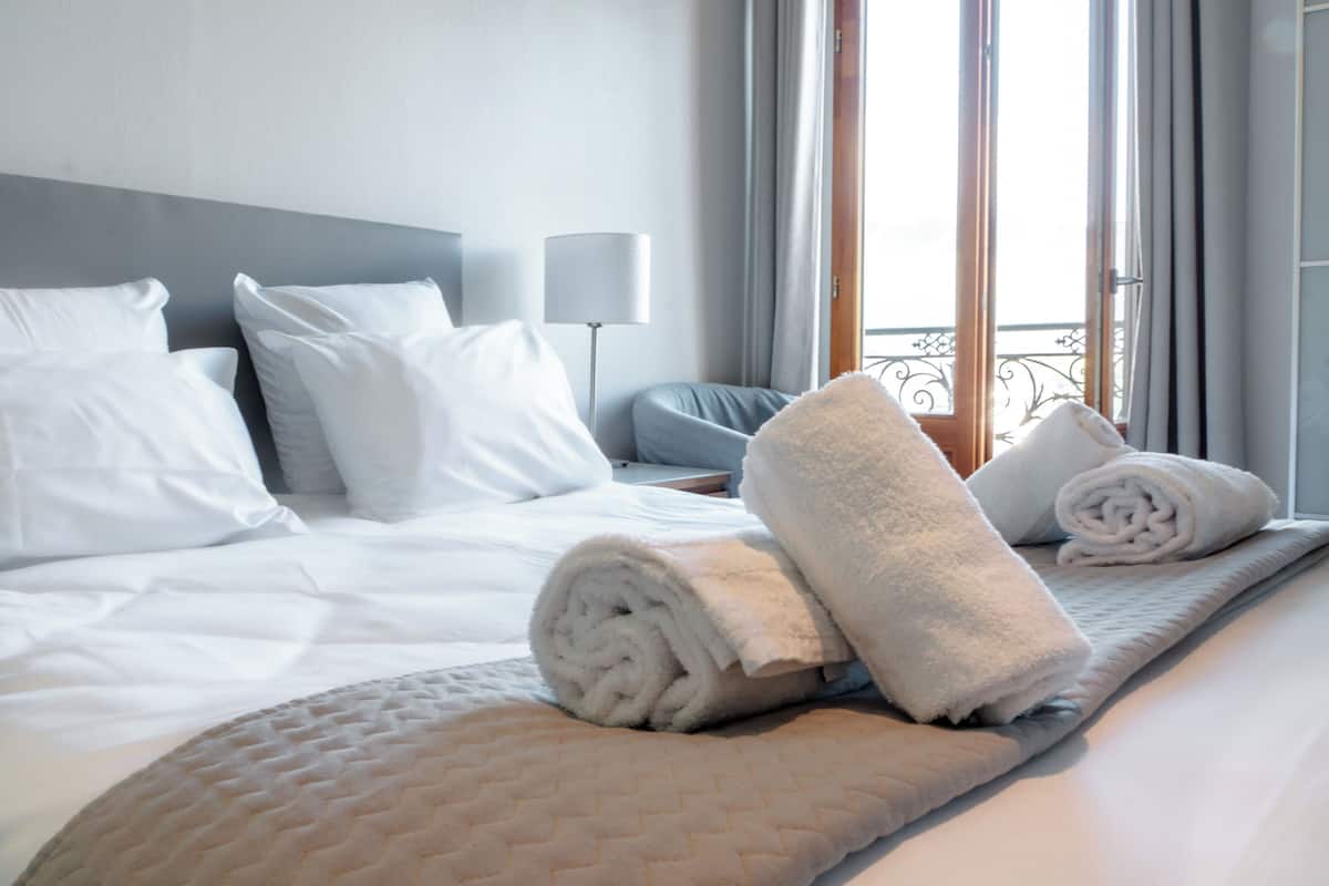 Habitación de hotel con cama, almohadas, una toalla enrollada, lámpara de noche y vistas al exterior.