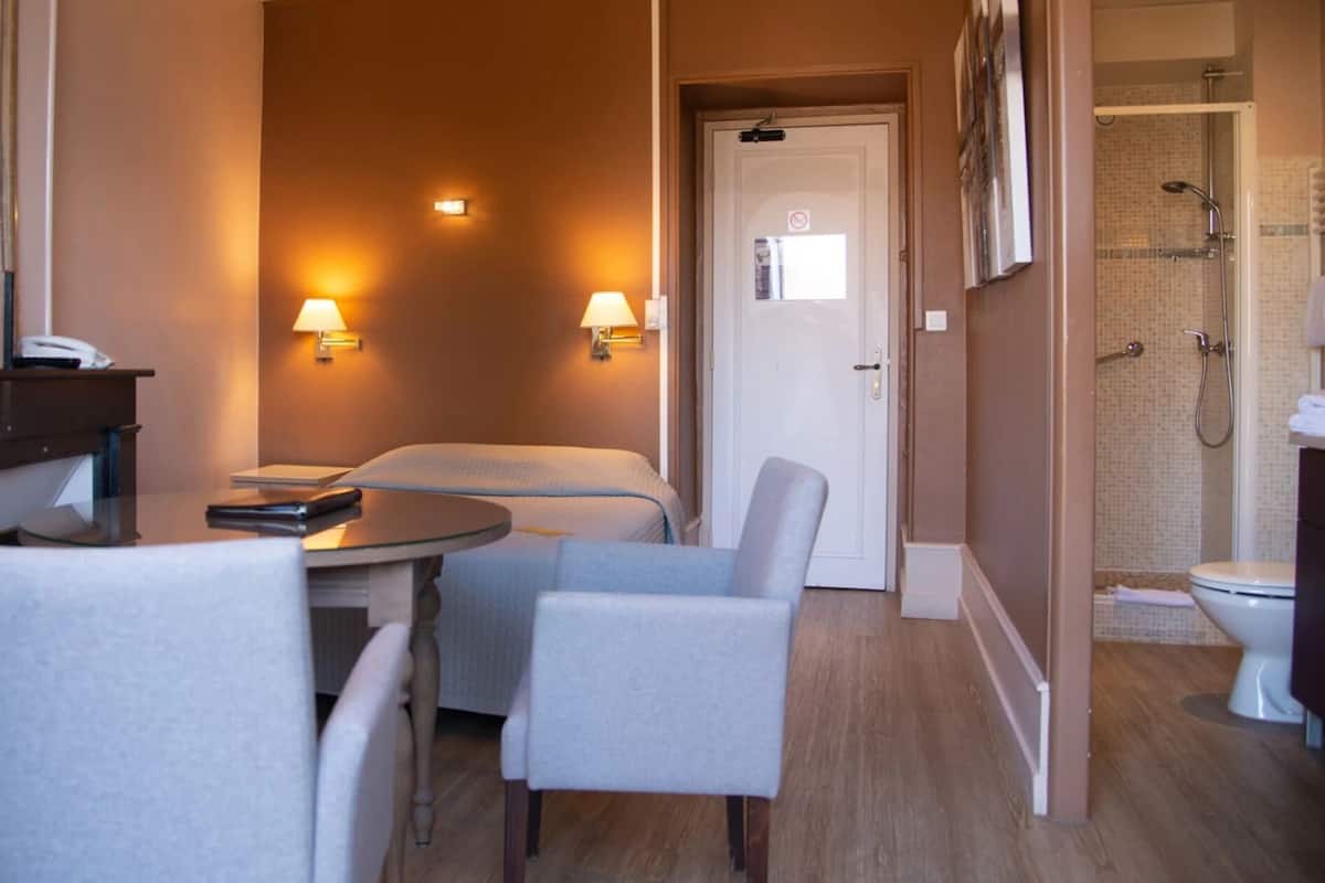 Habitación de hotel con cama, dos sillones, una mesita, un escritorio con teléfono, dos lámparas de pared, una puerta a zona de ducha y un inodoro.