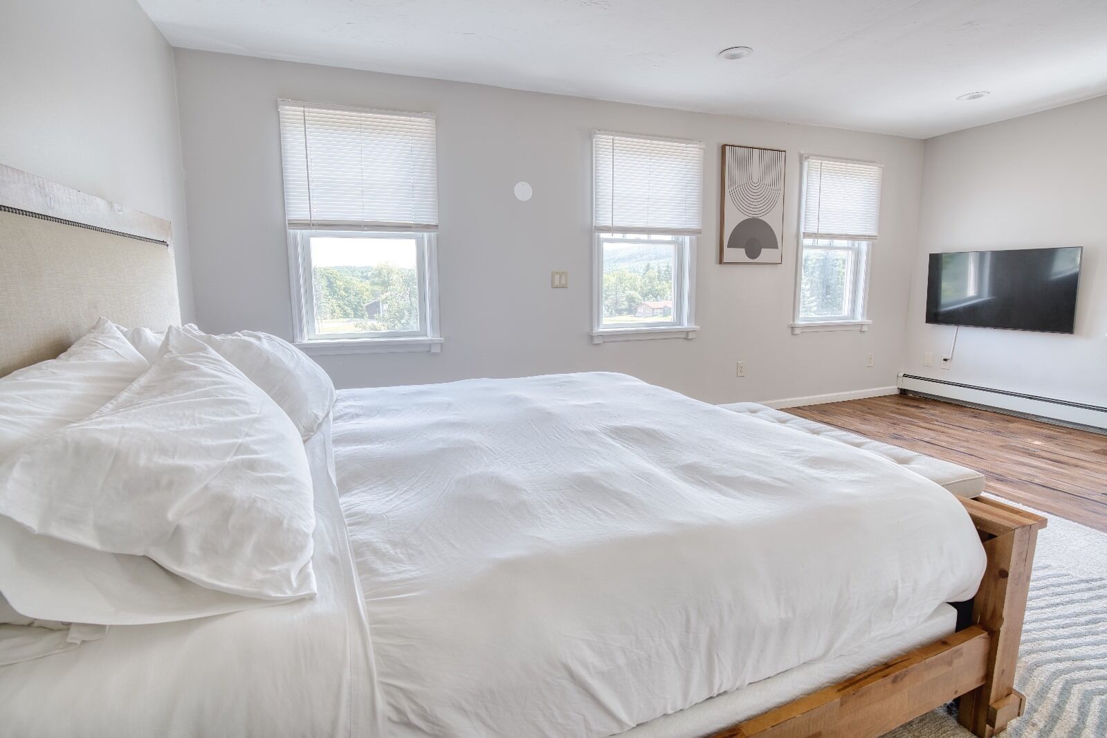Un dormitorio con una cama grande, dos ventanas con persianas, un televisor de pantalla plana en la pared y un cabecero de madera.