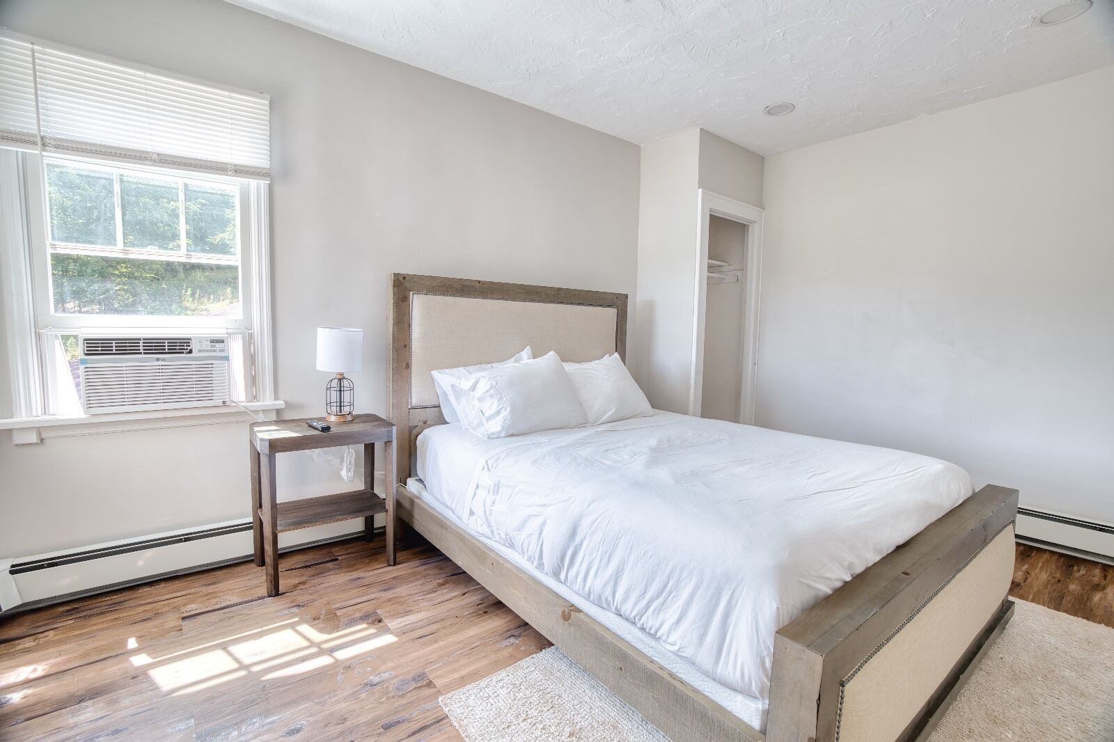 A bedroom with a wooden bed frame, white bedding, a bedside table with a lamp, and a window with blinds.