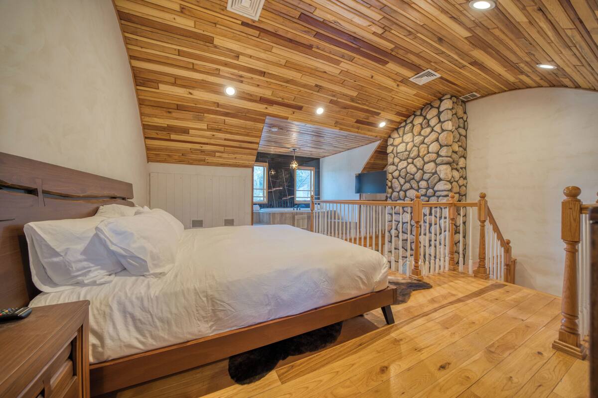 A bedroom with a wooden ceiling, a stone wall, a wooden bed, and a wooden staircase.