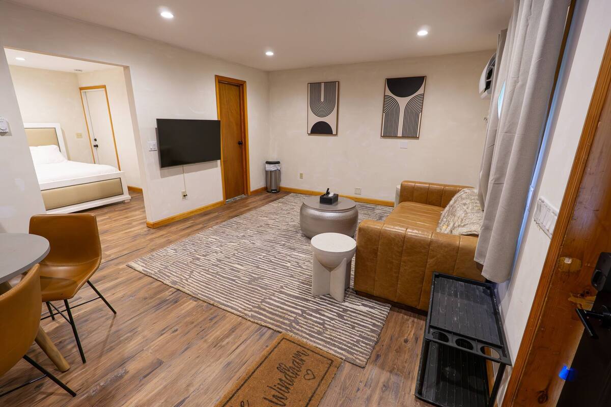 A modern hotel room with a bed, a sofa, a TV, a small table, and a trash bin.
