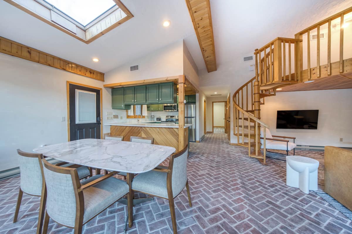 A spacious living area with a dining table and chairs, a kitchen with modern appliances, and a wooden staircase leading to an upper level.
