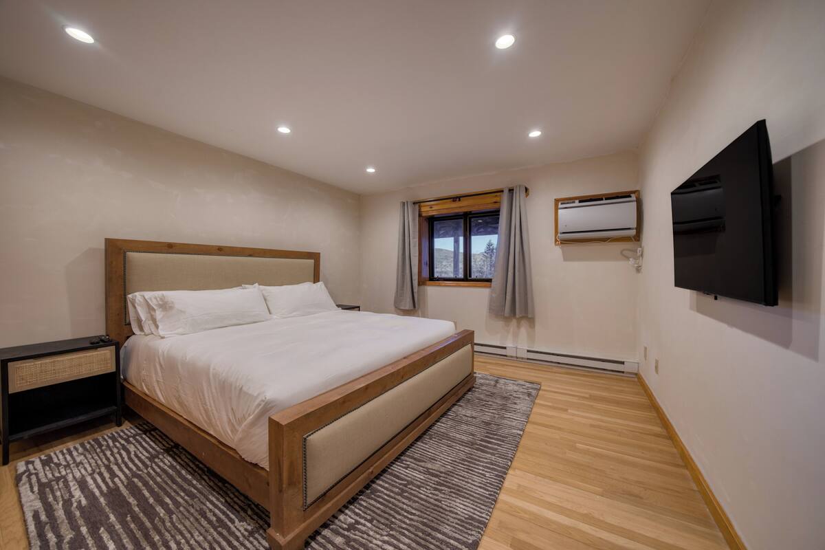 A bedroom with a large bed, a bedside table, a TV mounted on the wall, and a window with curtains.