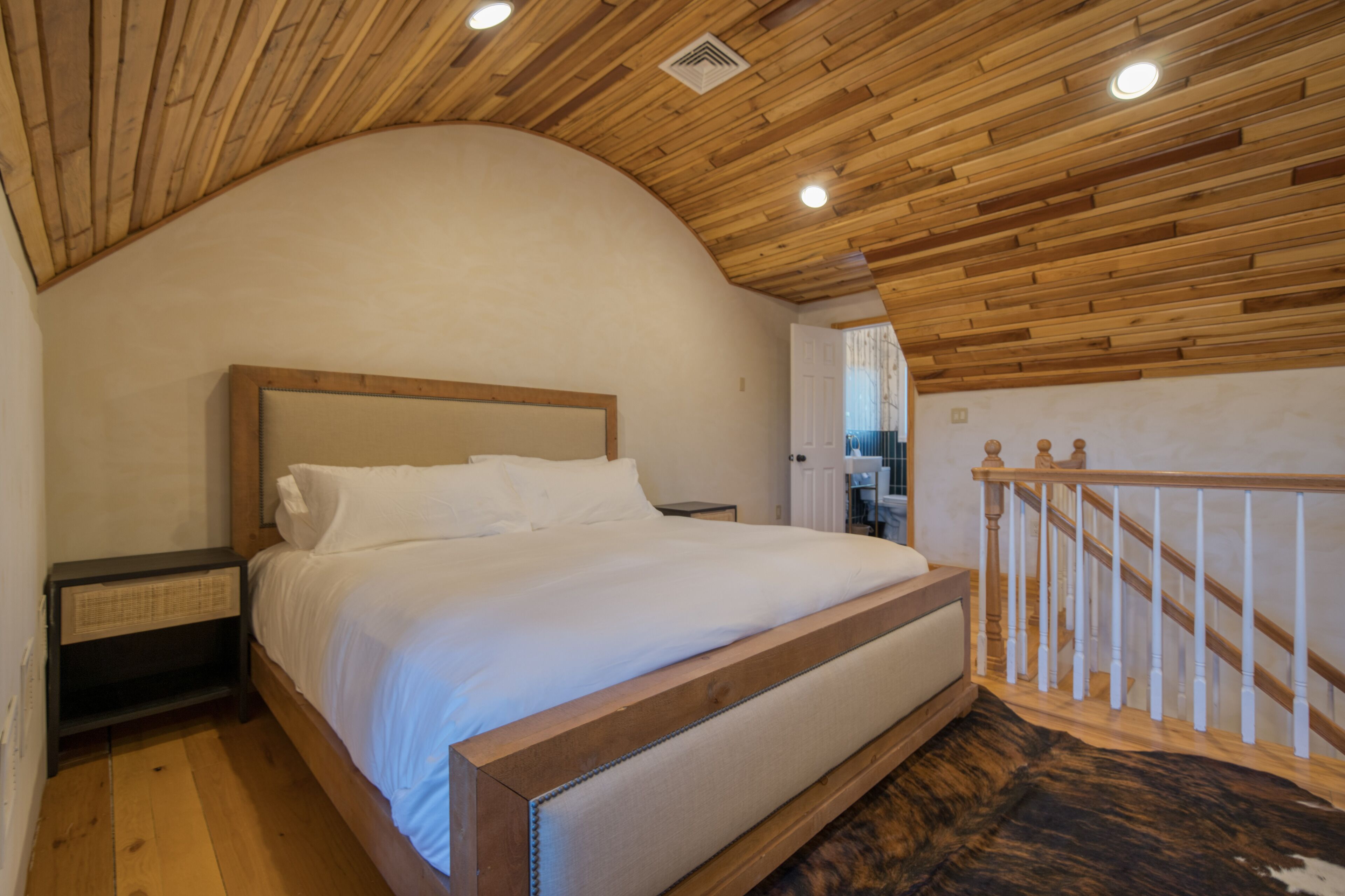 A bedroom with a wooden ceiling, a bed with white bedding, a nightstand, and a staircase with a wooden railing.
