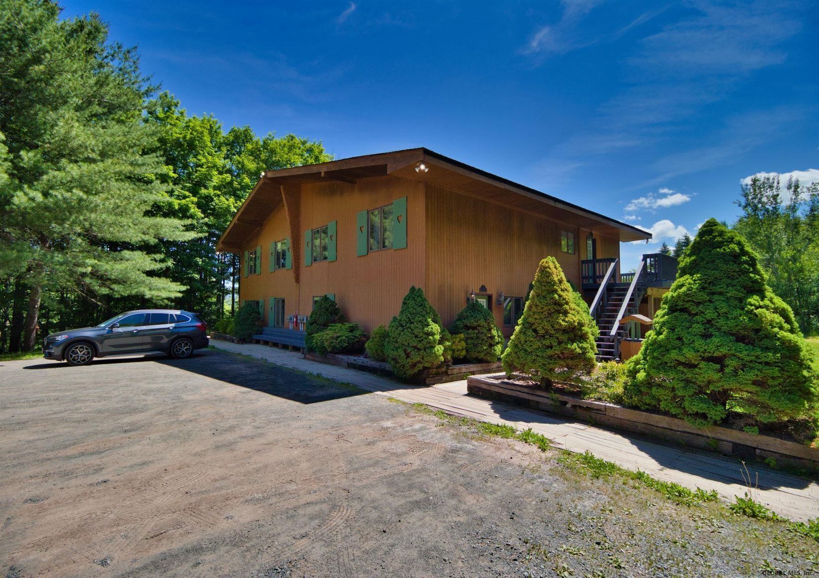 A two-story wooden building with a covered porch, surrounded by well-maintained greenery and a parked SUV.