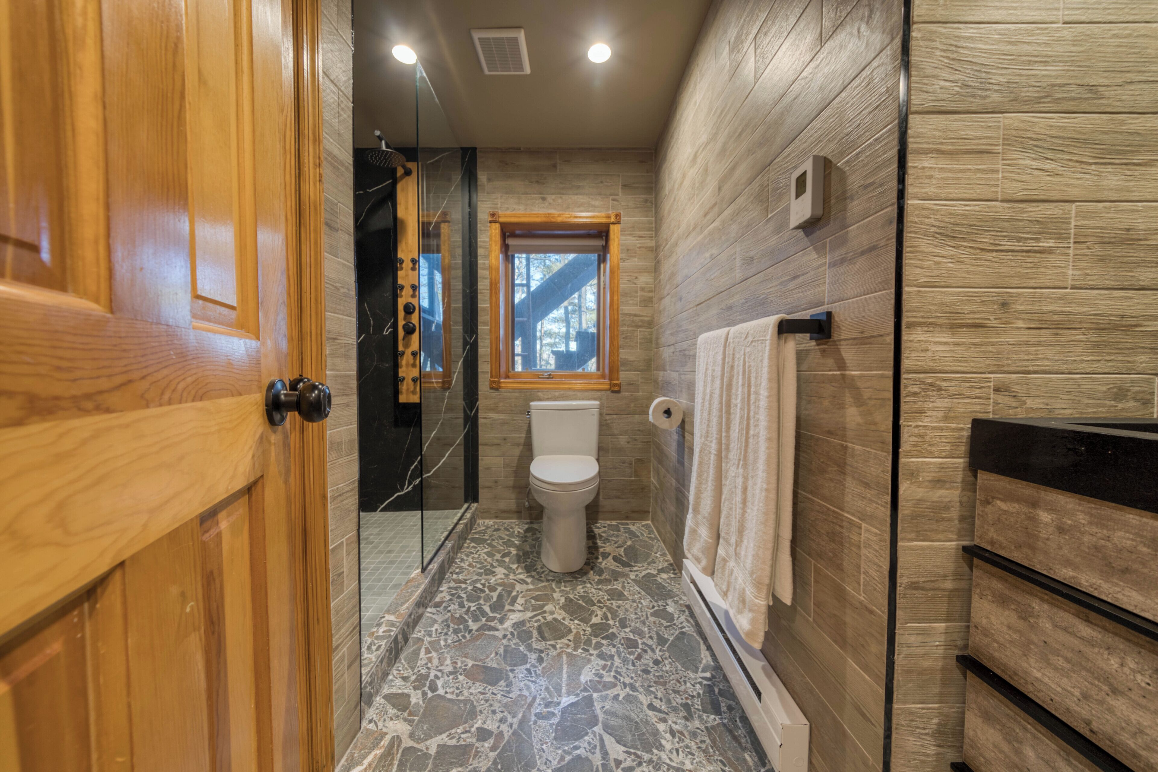 A bathroom with a wooden door, a toilet, a shower area, and a window.