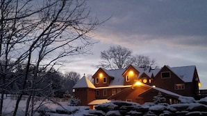 Una casa grande de madera de varios pisos con techo cubierto de nieve y varias ventanas iluminadas, rodeada de árboles desnudos y nieve.