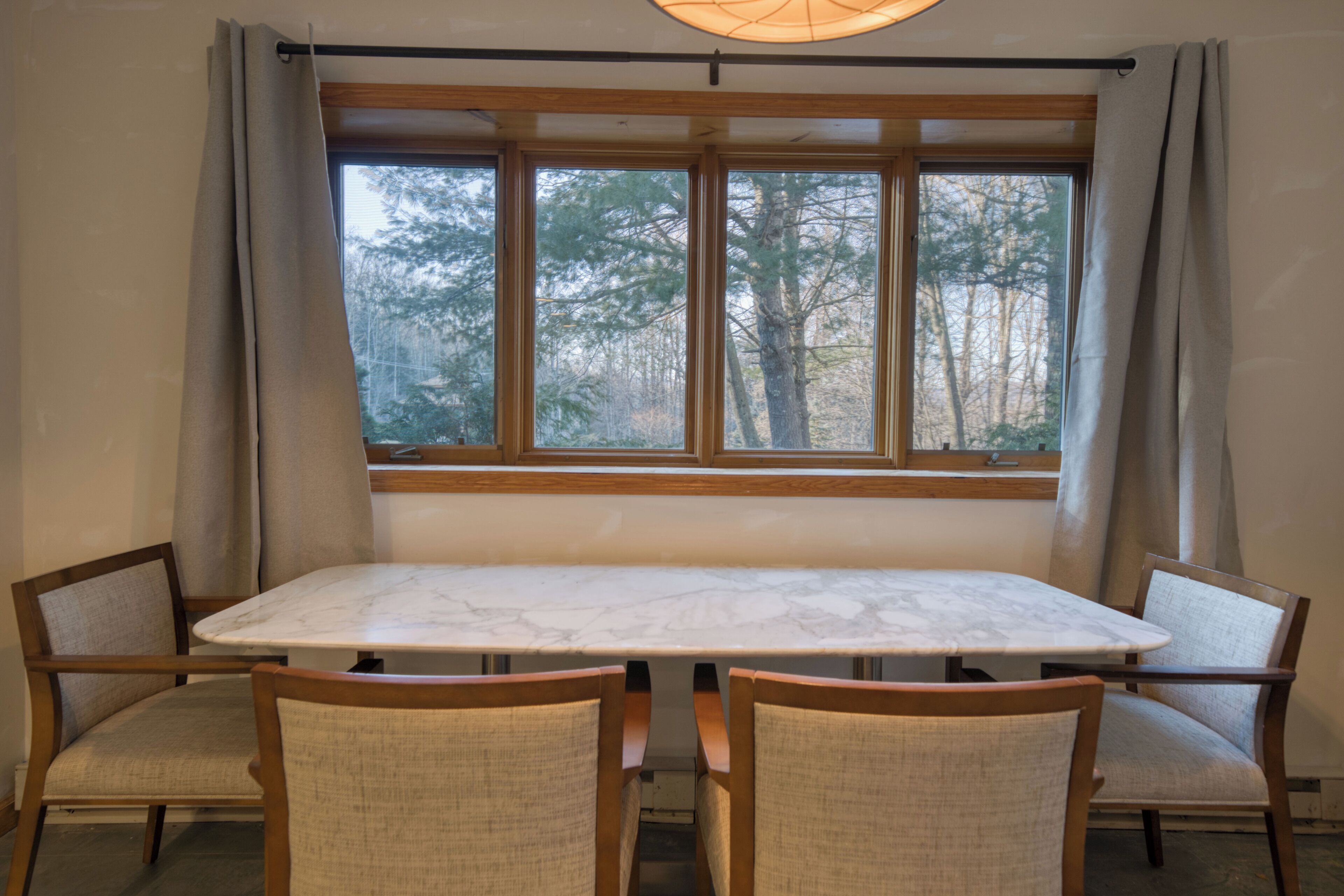 A dining area with a marble table and four chairs, a window with curtains, and a view of trees outside.