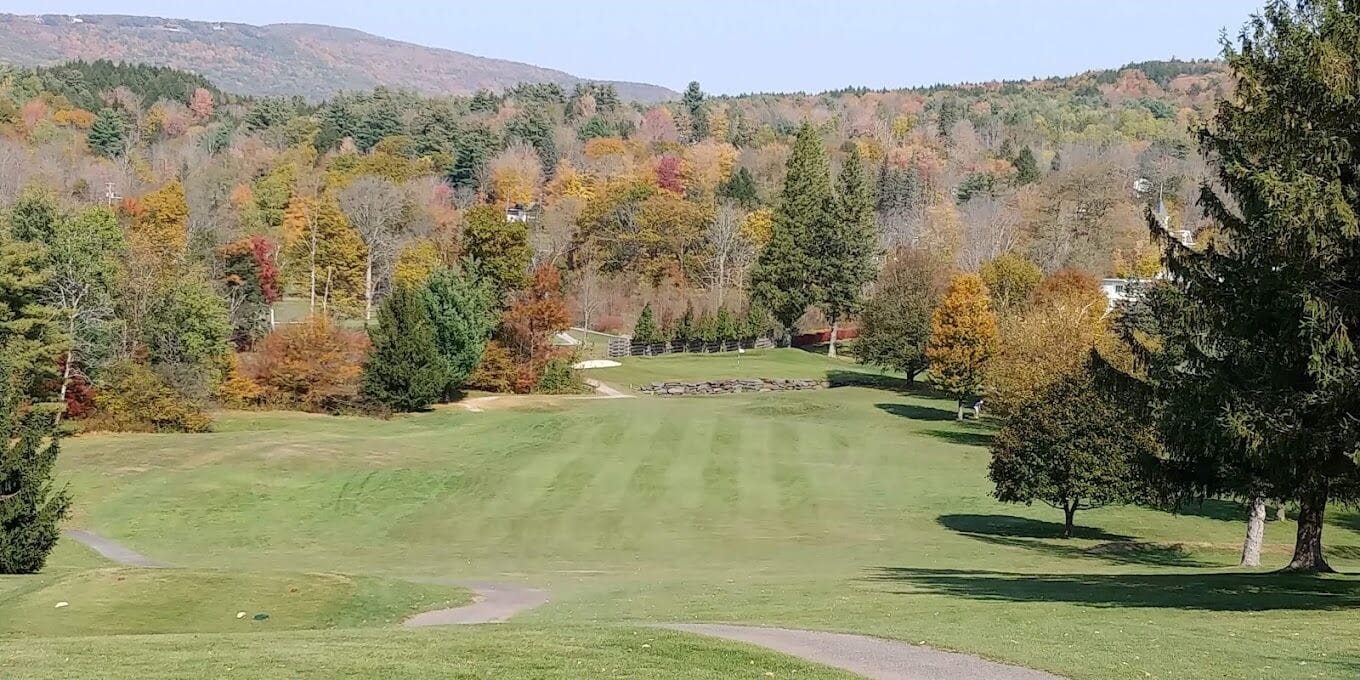 A golf course with a clear path, surrounded by trees with autumn foliage, and a hilly landscape in the background.
