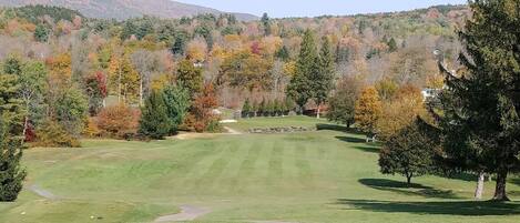 A golf course with a clear path, surrounded by trees with autumn foliage, and a hilly landscape in the background.