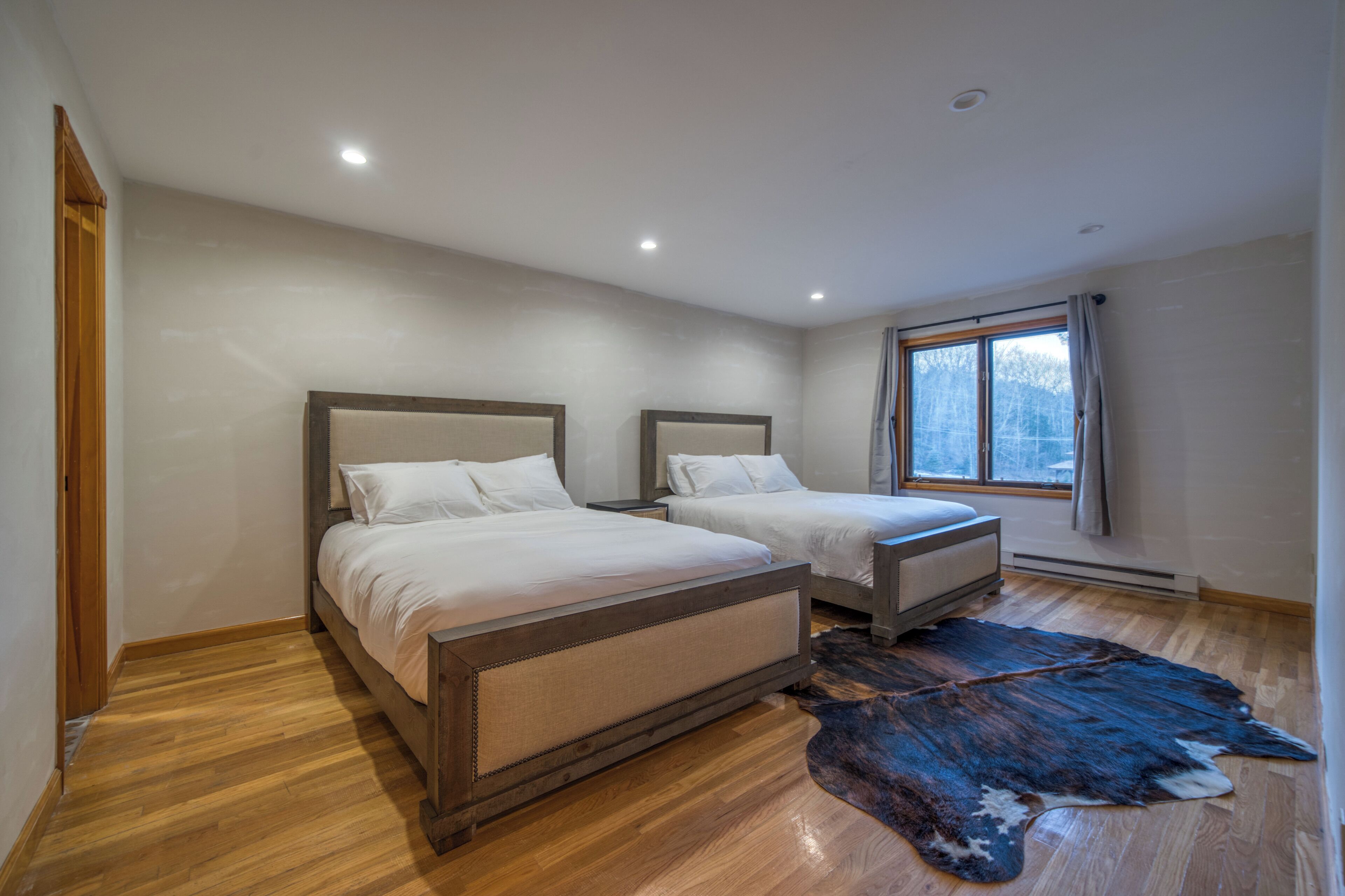 A bedroom with two beds, wooden flooring, a large window, and a fur rug.