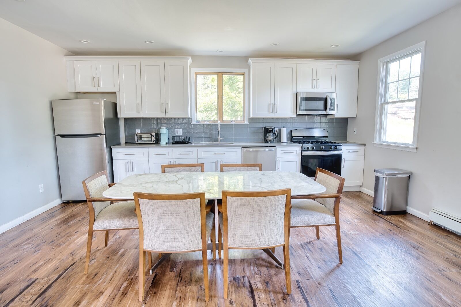 A modern kitchen with white cabinets, a stainless steel refrigerator, a microwave, and a stove. There is a dining table with chairs in the center.