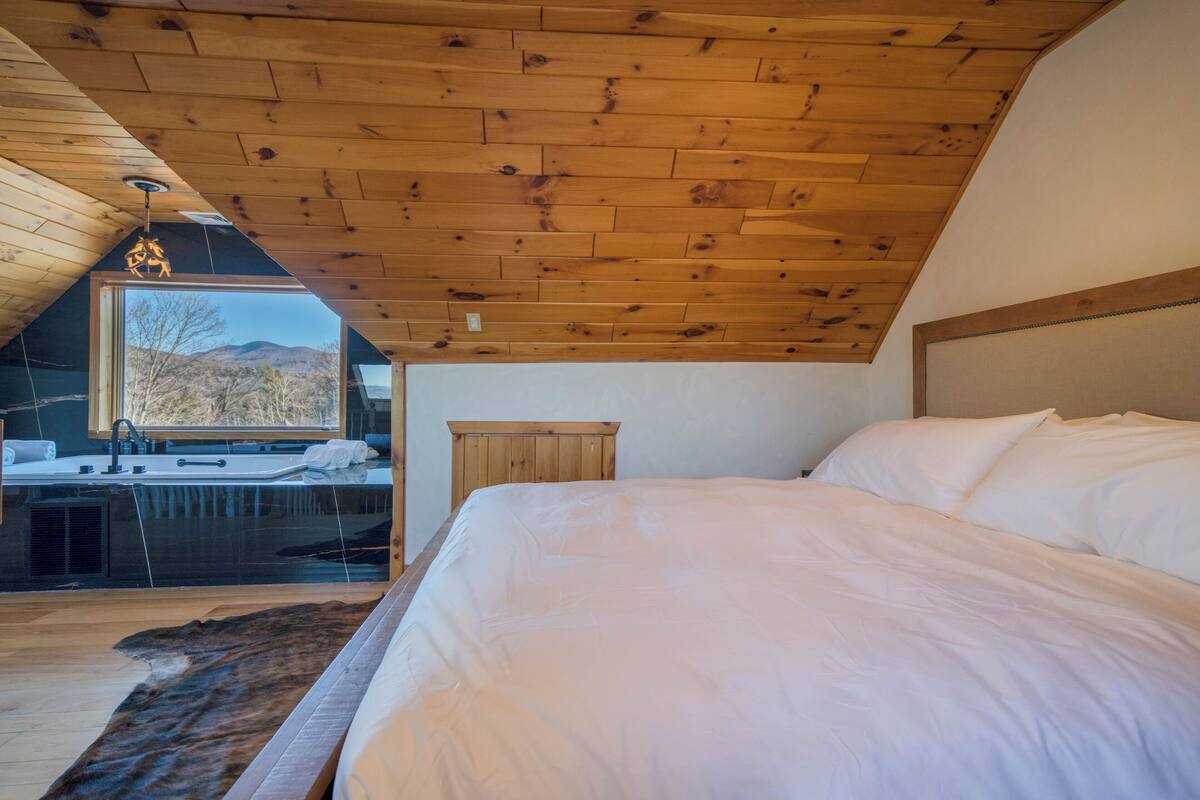 A bedroom with a wooden ceiling, a large bed, a window with a view of mountains, and a bathroom with a sink and mirror.