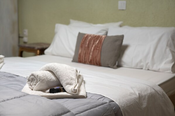 Una cama bien hecha con una toalla doblada y un libro, una mesita de noche de madera con una lámpara, y una pared verde.