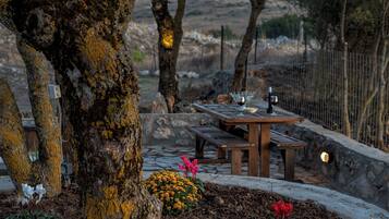 Un área de comedor al aire libre rústica con una mesa de piedra y bancos, rodeada de árboles y un muro de piedra.