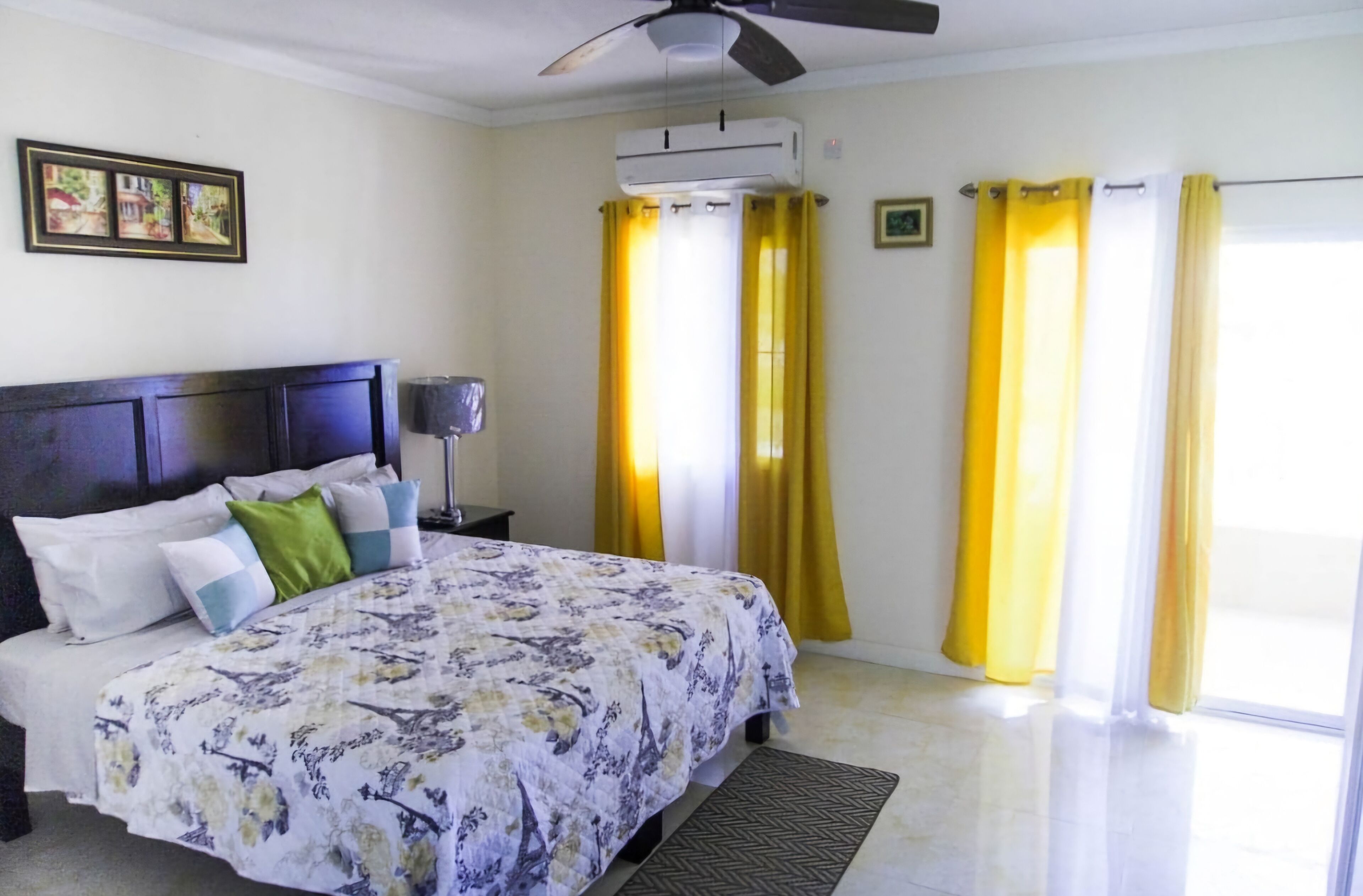 A bedroom with a bed, a ceiling fan, an air conditioning unit, and yellow curtains.