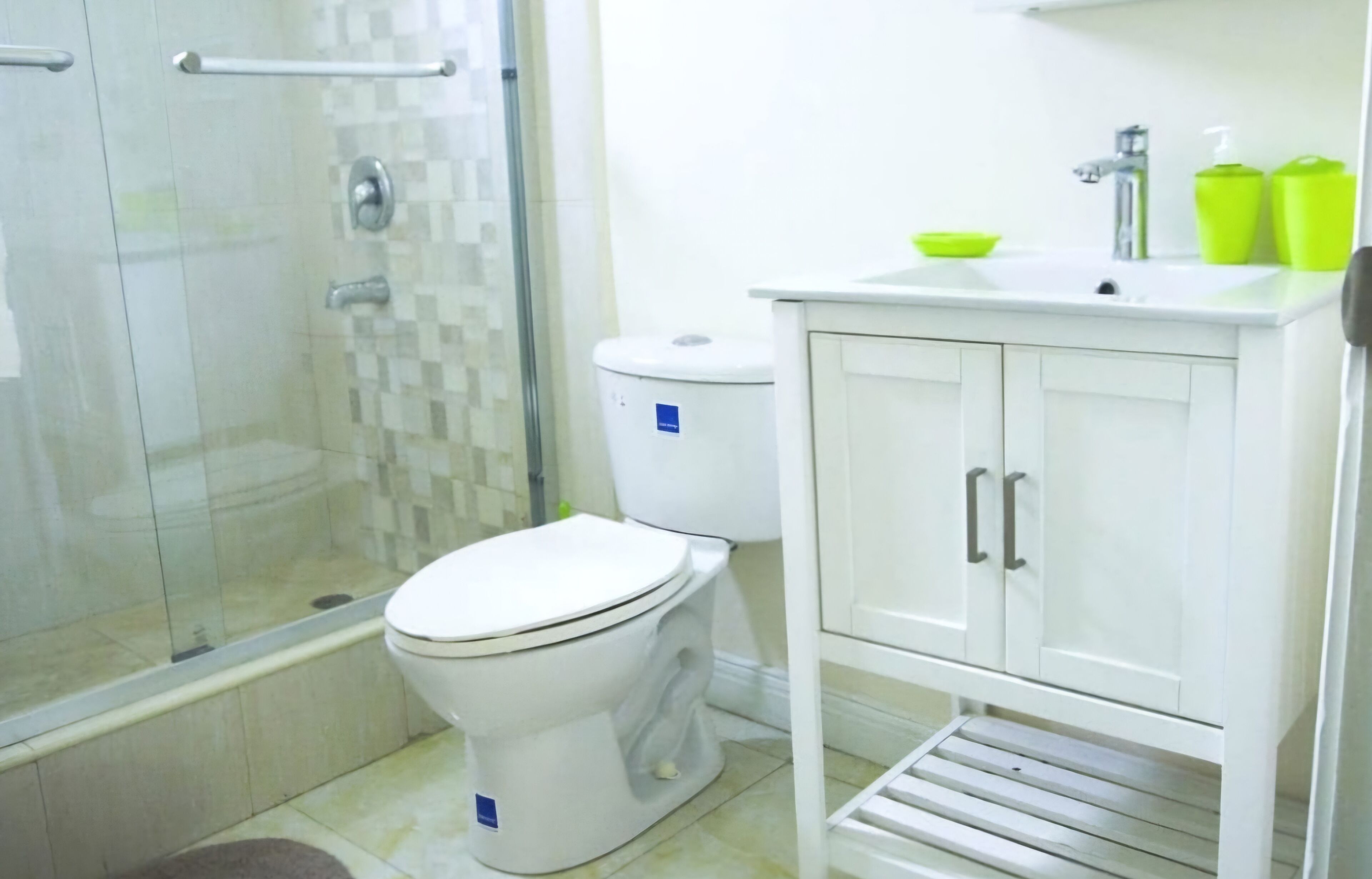 A bathroom with a glass shower enclosure, a white toilet, and a white sink with green soap dispensers.