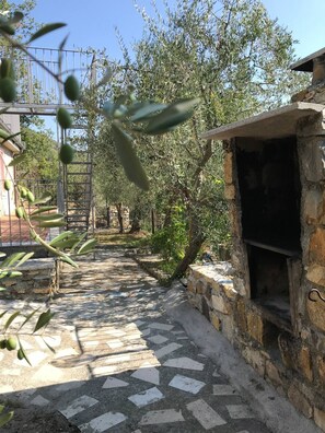 Un chemin de pierre mène à un espace extérieur rustique, agrémenté d’un barbecue en pierre, entouré d’oliviers et d’un portail en métal.