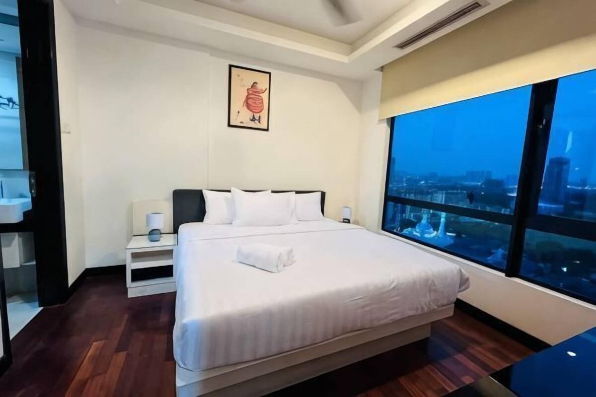 Una habitación de hotel con una cama grande, una mesita de noche, un ventanal con vistas a la ciudad y un cuadro enmarcado en la pared.