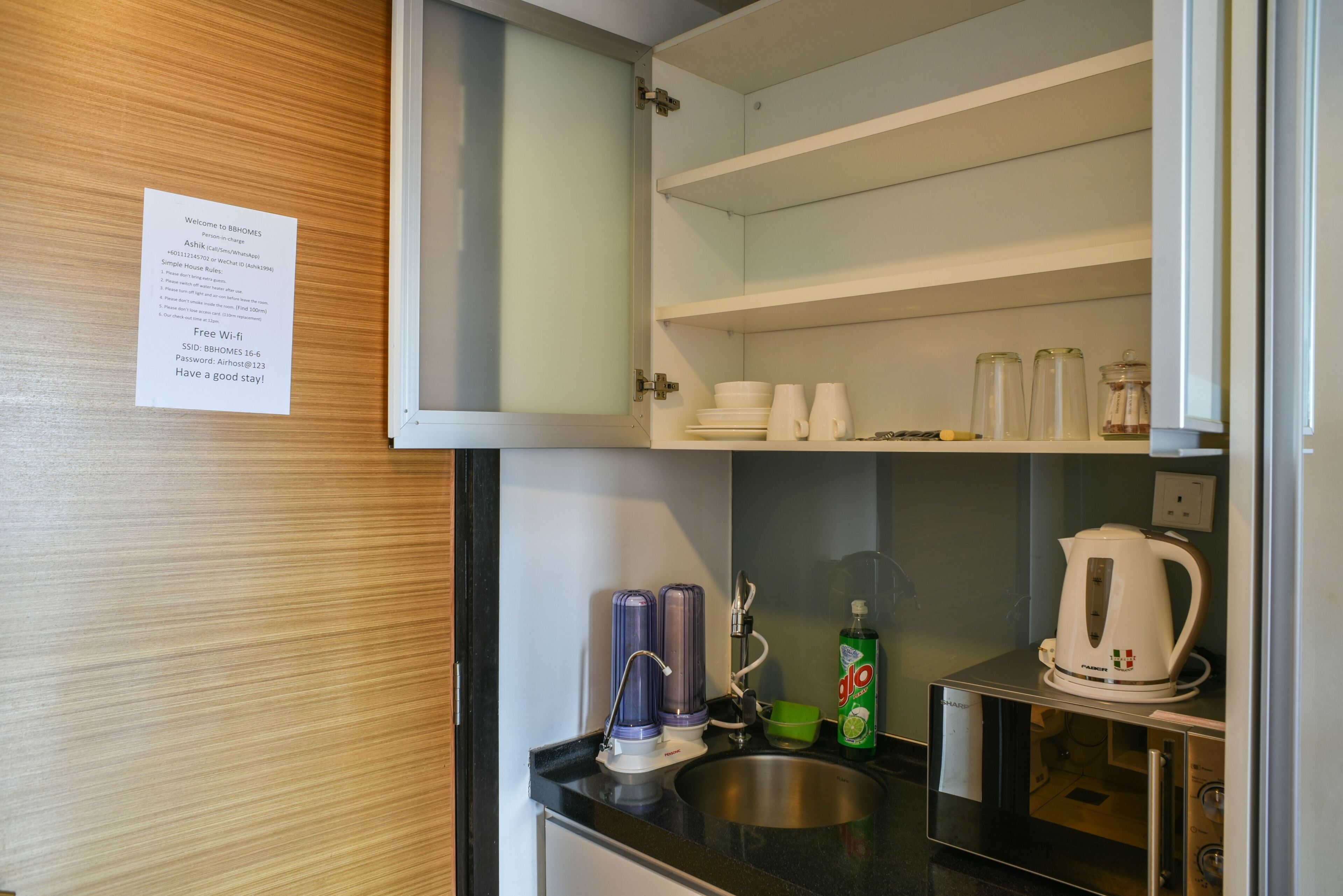 Een kleine keuken met een spoelbak, waterkoker en magnetron. Boven de spoelbak zijn planken en er hangt een briefje aan de muur.