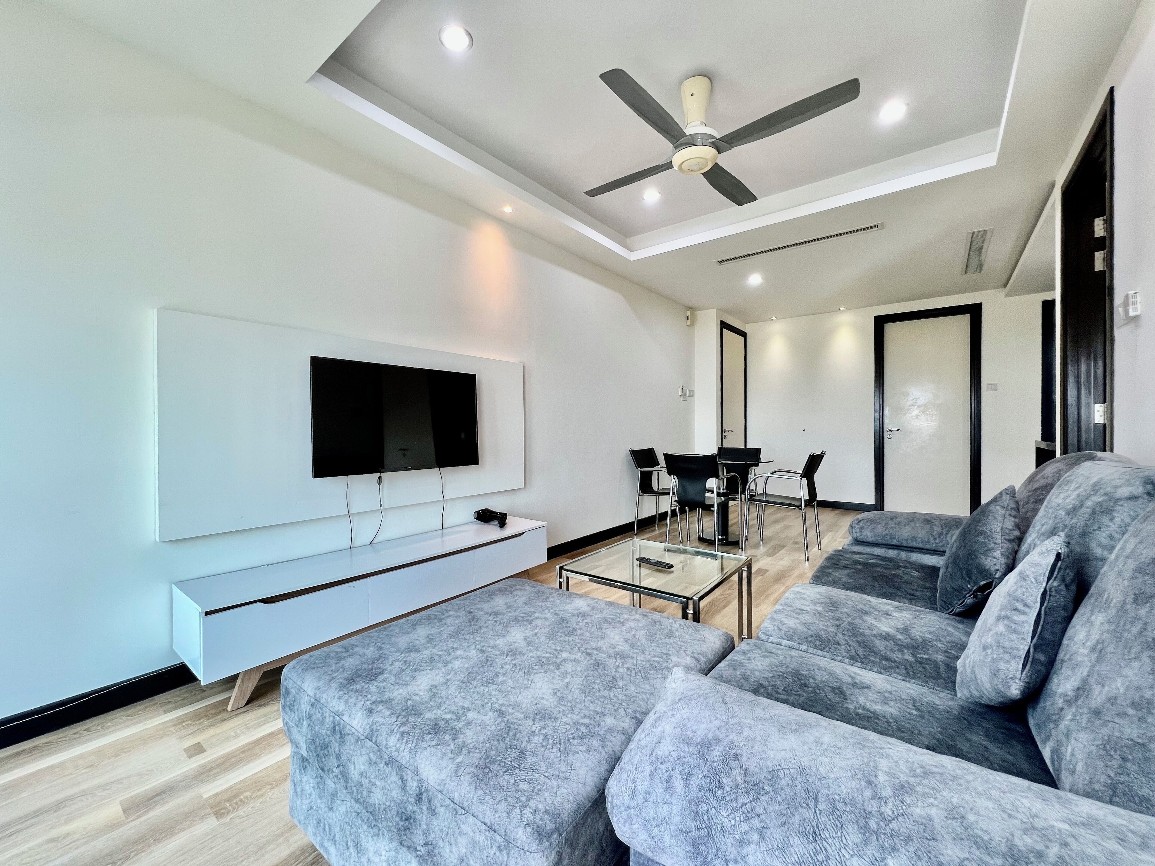 A modern living room with a flat-screen TV, grey sofa, ceiling fan, and a dining area with a table and chairs.