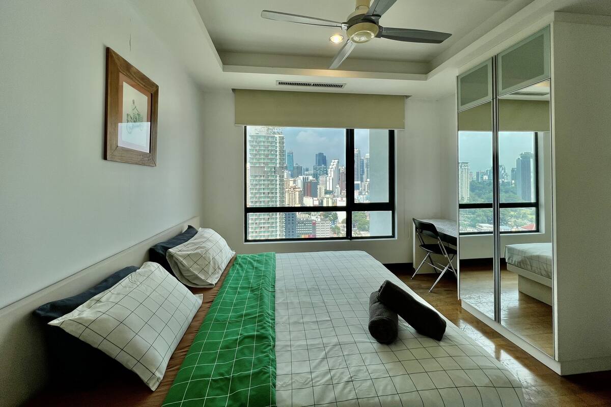 Un dormitorio con cama, ventilador de techo, ventana grande con vista a la ciudad y un cuadro en la pared.