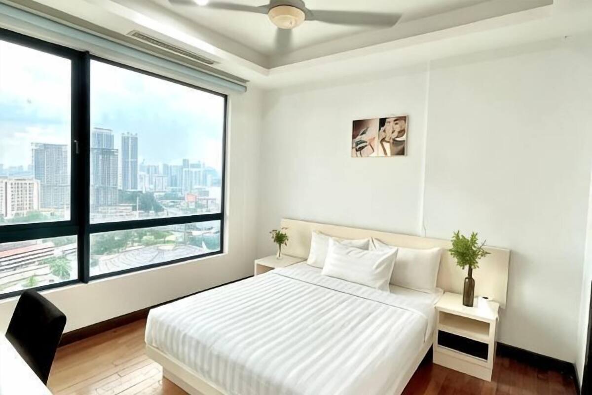 Un dormitorio con una cama grande, una mesita de noche, una silla, un ventanal con vista a la ciudad y un ventilador de techo.