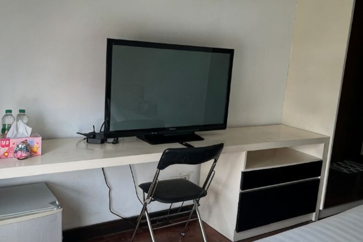 Una habitación con televisión, escritorio, silla y un pequeño armario.