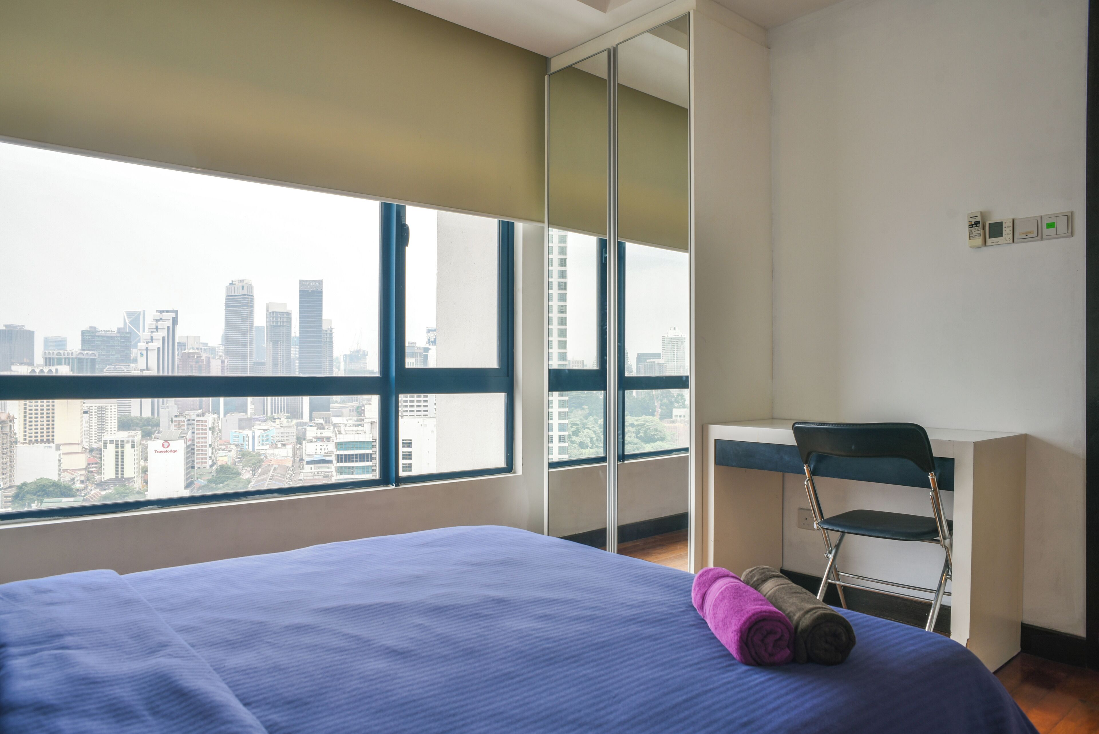 Ein Hotelzimmer mit einem großen Bett, einem Schreibtisch und einem Stuhl. Durch große Fenster bietet das Zimmer Blick auf die Stadt.