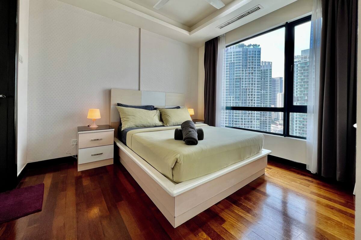 Una habitación de hotel moderna con una cama grande, una mesita de noche con lámpara, un ventilador de techo y vistas a edificios altos por la ventana.