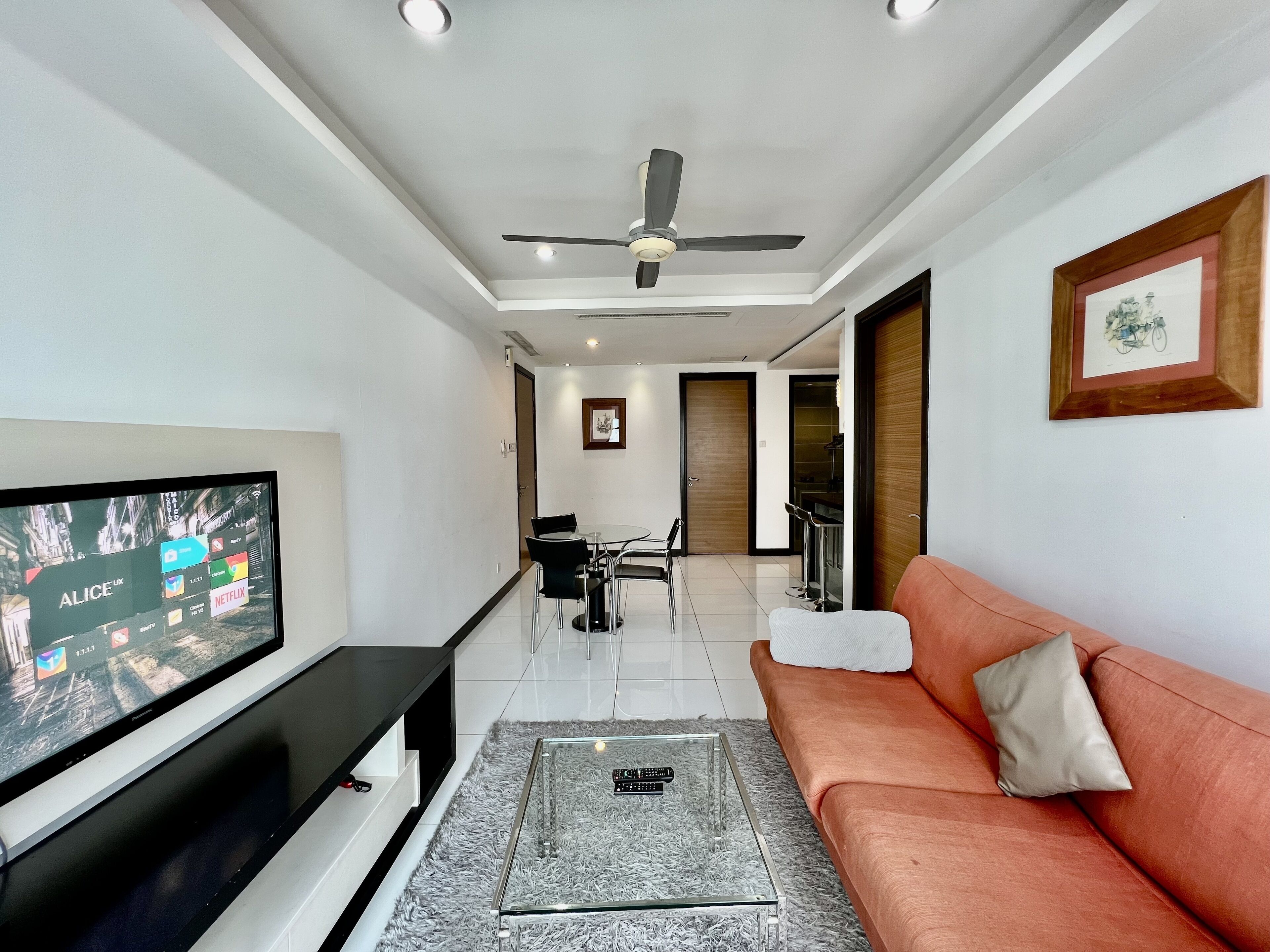 A modern living room with a flat-screen TV, a glass coffee table, a sofa, and a ceiling fan.