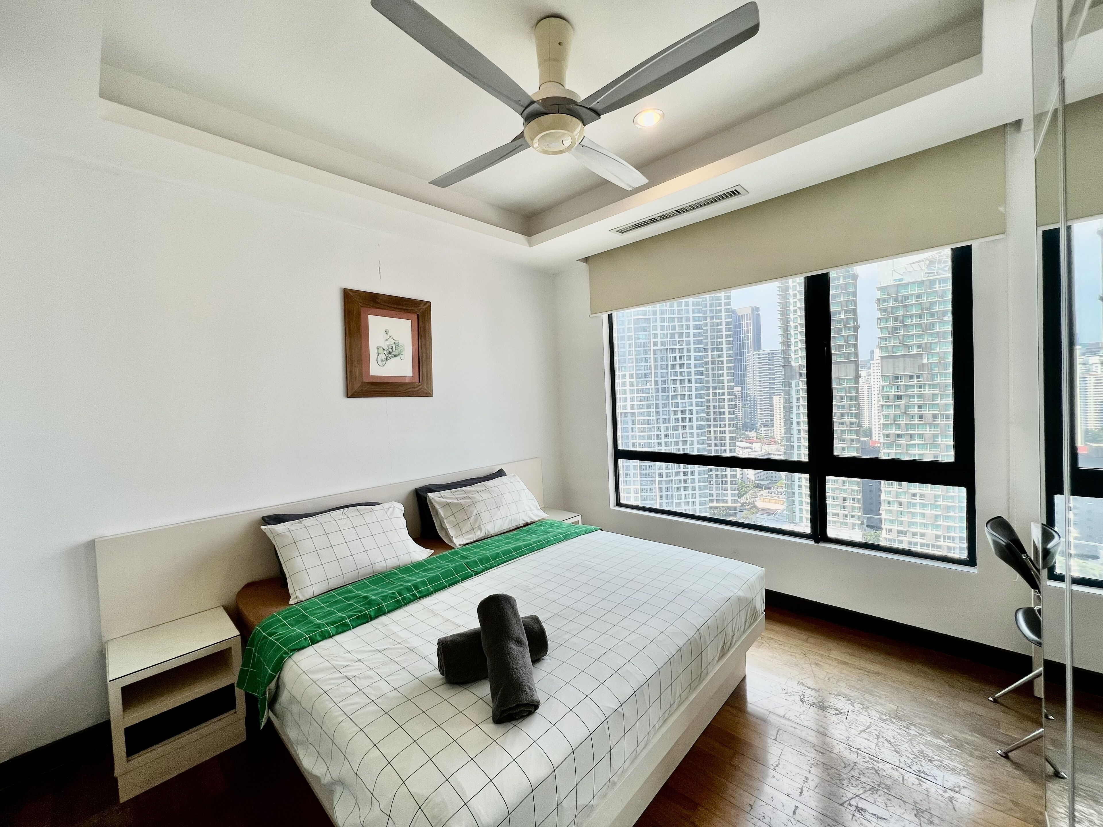 Un dormitorio con cama, ventilador de techo, ventana con vista a la ciudad y un cuadro en la pared.