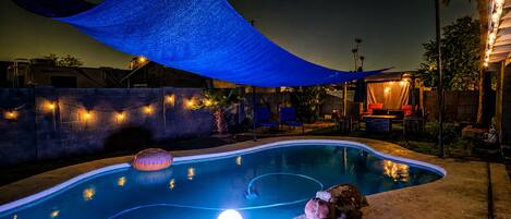 A swimming pool with a blue shade sail overhead, a lounge area with chairs, and a small water feature.