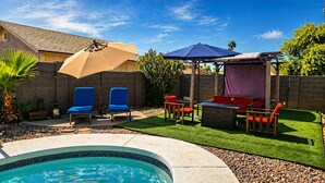 Ein Garten mit Swimmingpool, blauen und roten Liegestühlen, einer Pergola mit Dach und einem gepflegten Rasen.