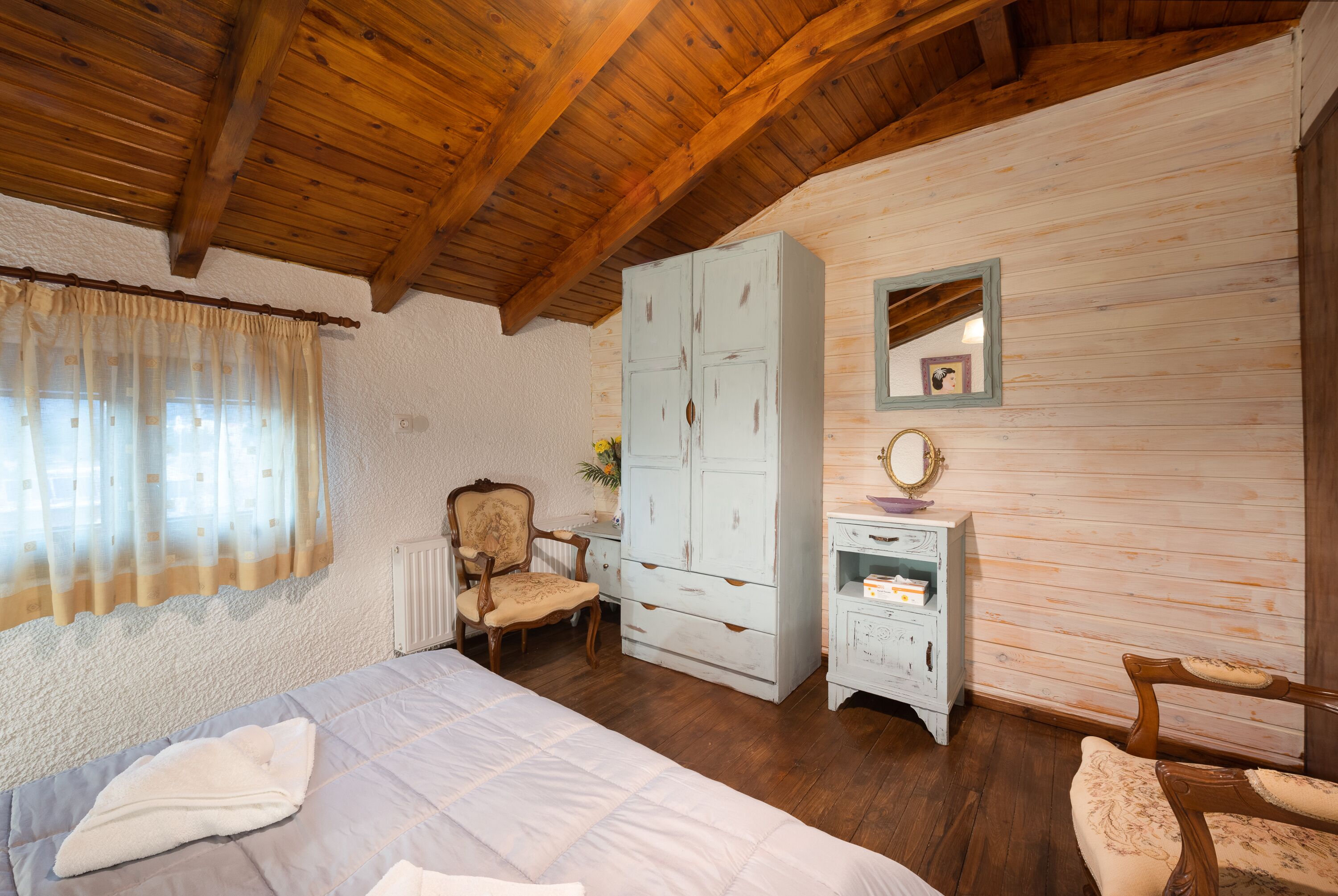 A bedroom with a wooden ceiling, a bed with a white duvet, a wardrobe, a chair, and a small bedside table with a mirror.