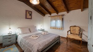 Ein Schlafzimmer mit Holzbalkendecke, einem Bett mit beiger Bettdecke, zwei Nachttischen mit Lampen, einem Sessel und einem Fenster mit Vorhängen.