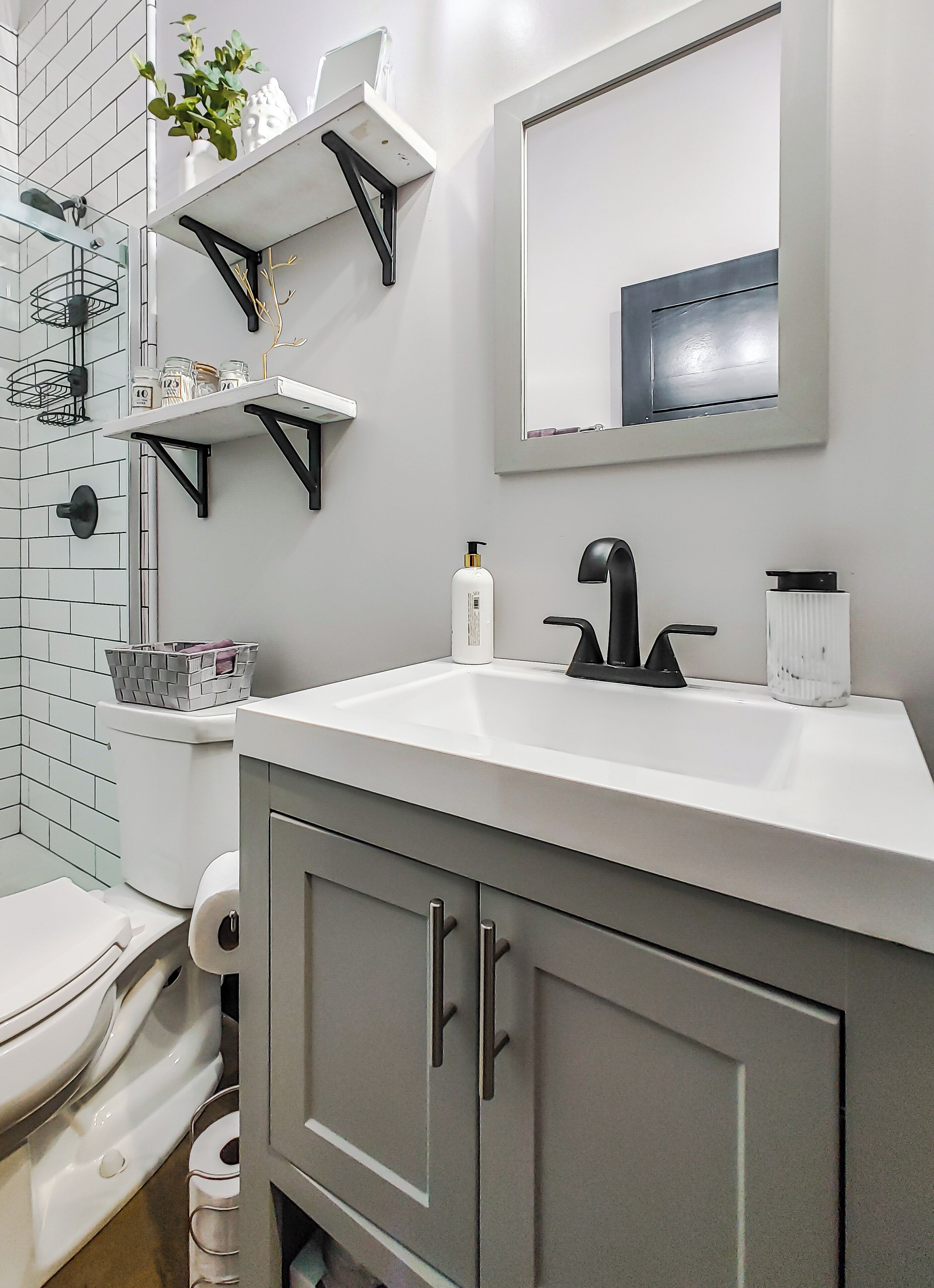 A modern bathroom with a white sink, black faucet, and grey cabinets. There are shelves with toiletries and a mirror above the sink.
