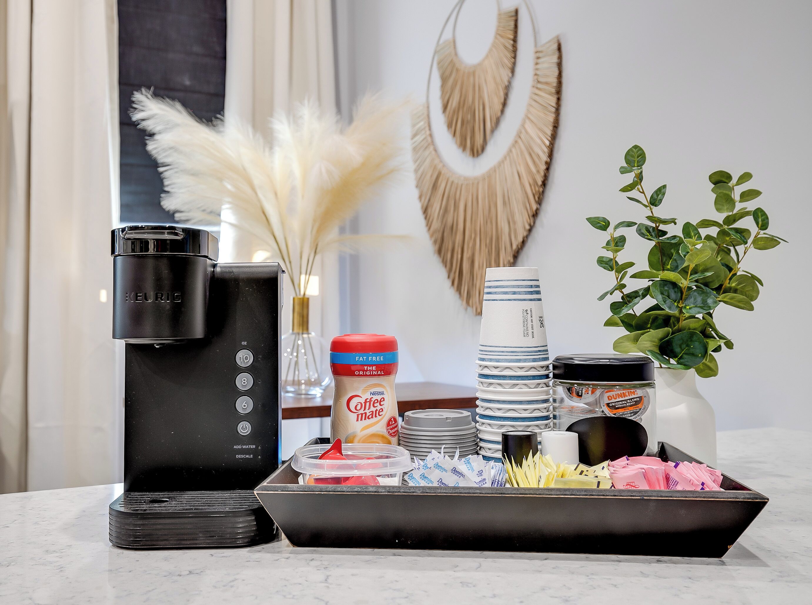 Una encimera de cocina con una cafetera Keurig, una taza de café, un dispensador de cápsulas de café, una pila de vasos desechables y una bandeja de cápsulas.