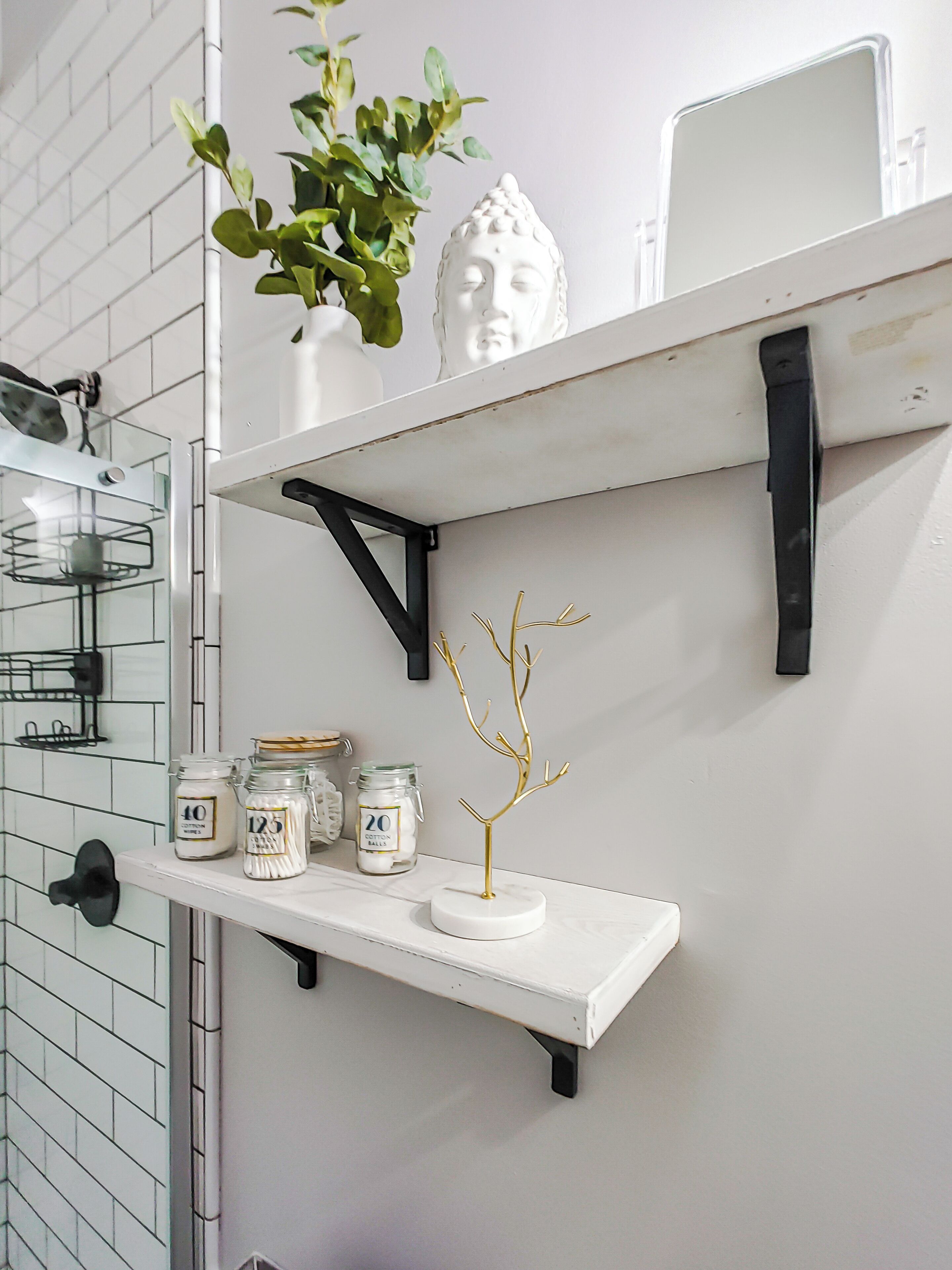 A bathroom with white tiled walls, a white shelf with jars, a white Buddha statue, and a potted plant.