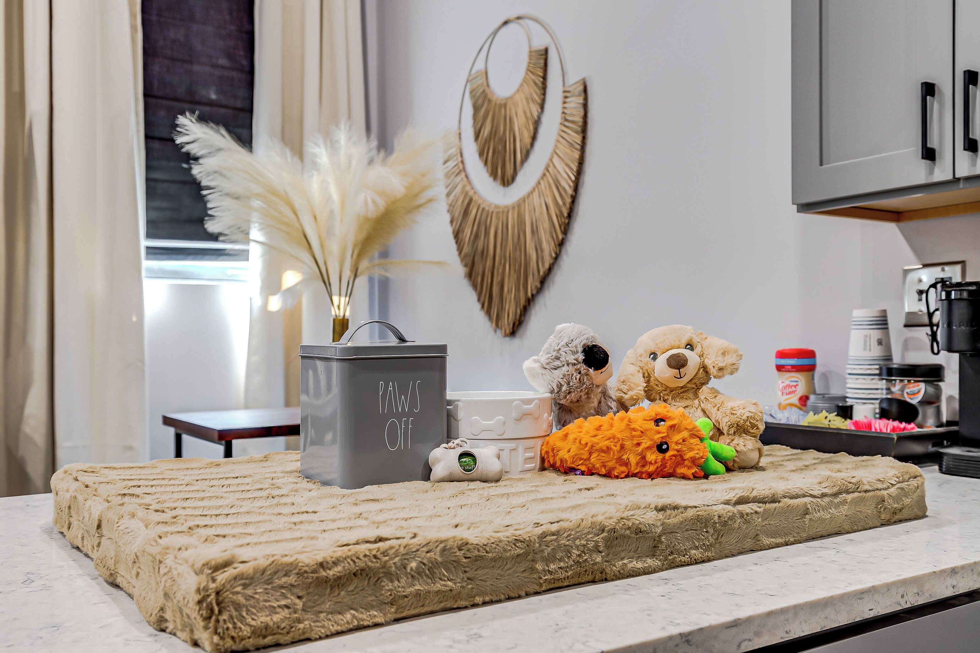 Una cocina con encimera de mármol, un cubo de basura gris con la etiqueta 'Paws Off', un jarrón con pampas secas y dos osos de peluche sobre una alfombra beige.