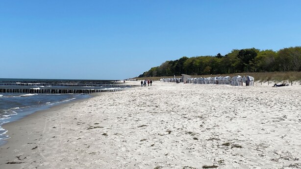 A sandy beach with a wooden pier extending into the water, a row of beach huts, and a forested hill in the background.