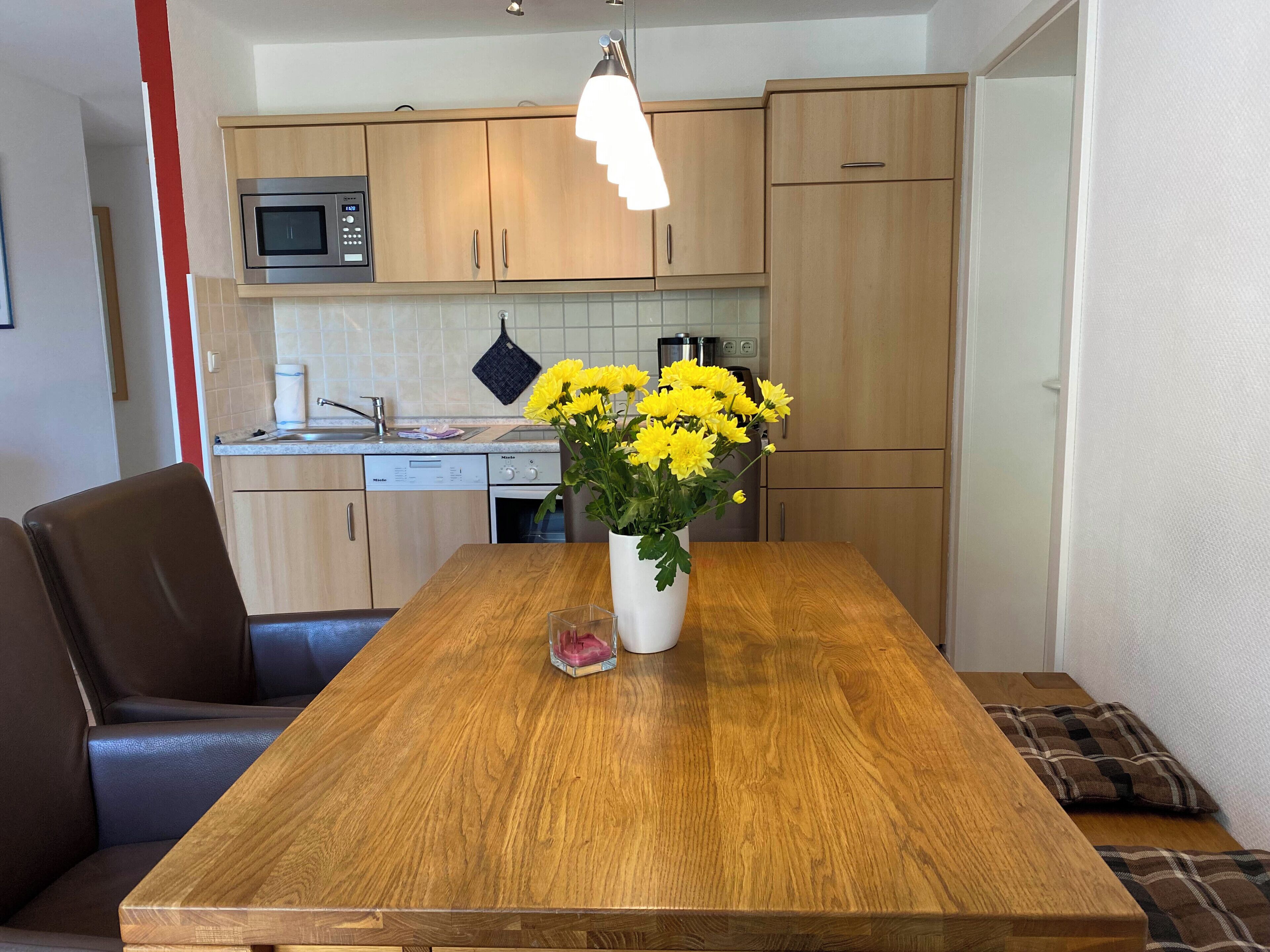 A modern kitchen with wooden cabinets, a built-in microwave, and a dining table with a vase of yellow flowers.