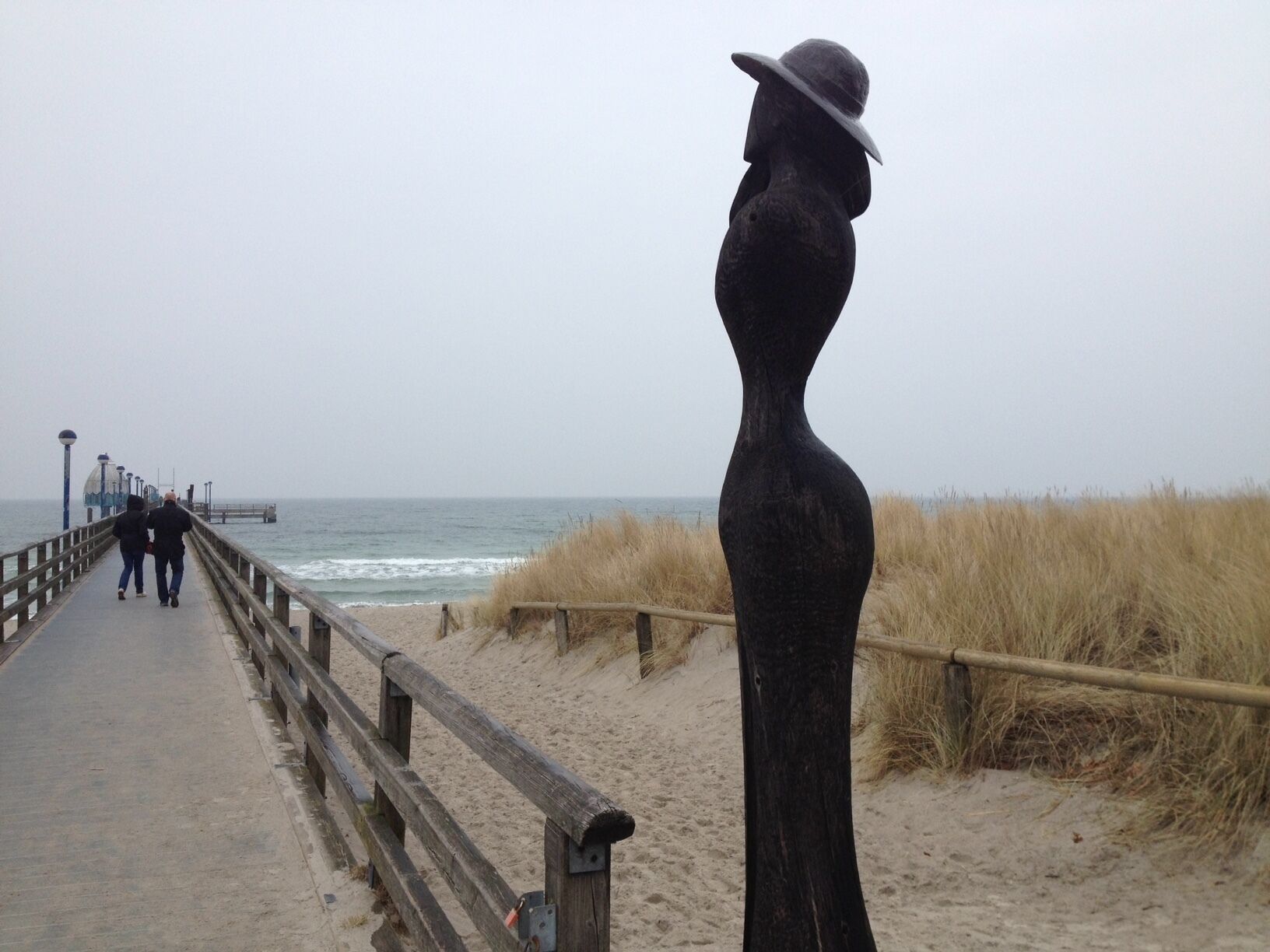 A boardwalk with a sculpture of a woman in a hat, a wooden railing, and the ocean in the background.
