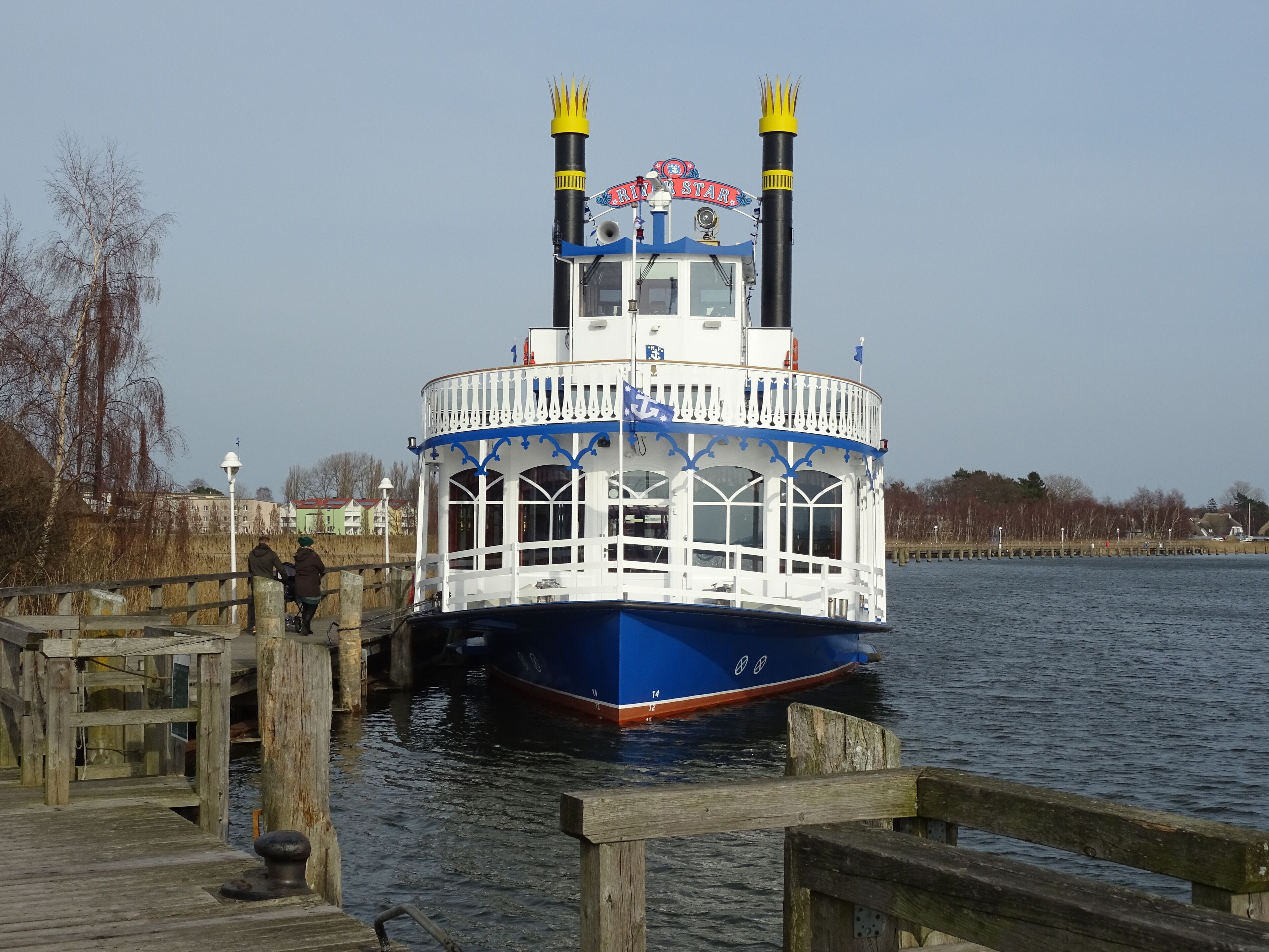Un bateau fluvial était amarré à un quai. Il était peint en blanc et bleu, possédait une cabine avec des fenêtres et un drapeau flottait au sommet.