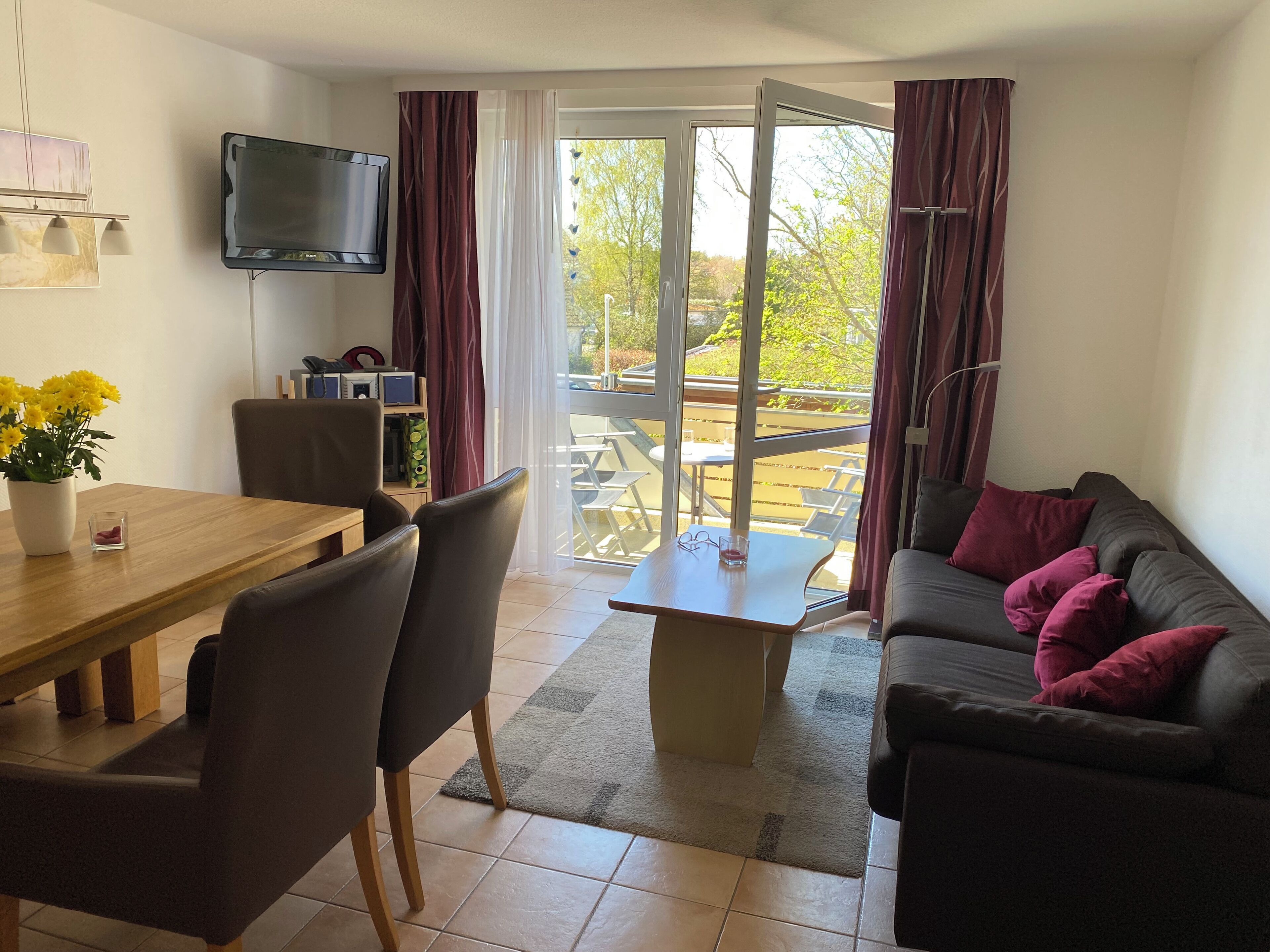 Een woonkamer met een bank, een eettafel met stoelen, een aan de muur bevestigde televisie en uitzicht buiten door een glazen deur.